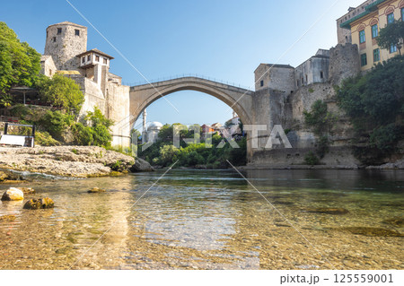 The Stari Most bridge, old bridge on Neretva river in Mostar town, Bosnia and Herzegovina, Europe. Arching stone bridge spans a tranquil river, connecting ancient structures under a clear sky. 125559001