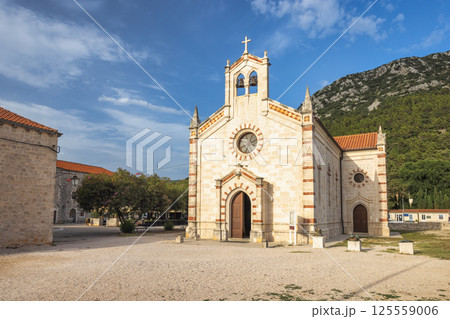 Ston, historic town in Peljesac peninsula in Croatia, Europe. Picturesque church with stone facade and cross-topped bell tower against blue sky near lush mountain landscape. Ston, historic town in Peljesac peninsula in Croatia, Europe. Picturesque church with stone facade and cross-topped bell tower against blue sky near lush mountain landscape. 125559006