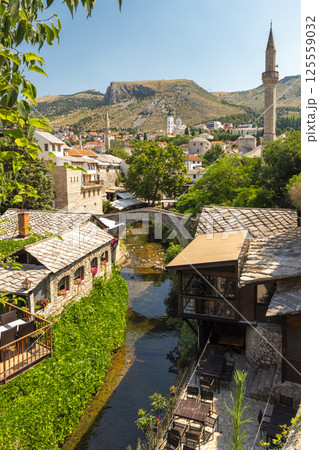 Mostar, historic town in Bosnia and Herzegovina, Europe. Charming riverside town with stone buildings, a bridge, lush greenery, and a minaret against a backdrop of hills and a clear blue sky. 125559032