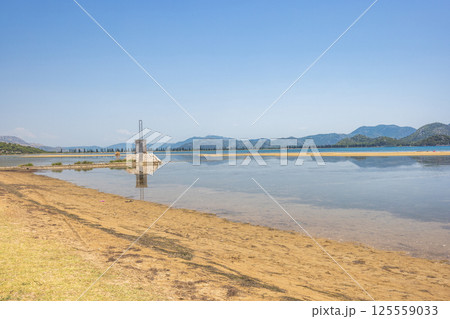 Delta of the Neretva river near the mouth of river into Adriatic Sea, Croatia, Europe. Scenic landscape featuring a calm coastline, a small building in water, and distant mountains beneath a blue sky. 125559033