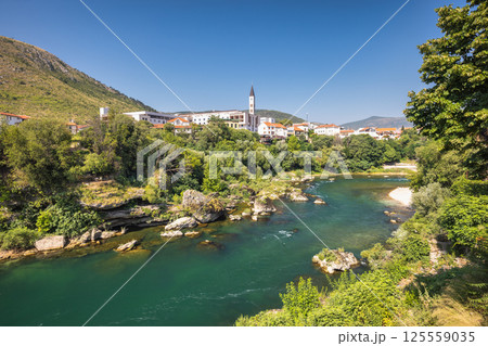 Neretva river in Mostar town, Bosnia and Herzegovina, Europe. River flows through a European town. Lush greenery surrounds buildings with mountains in background under a bright blue sky. Neretva river in Mostar town, Bosnia and Herzegovina, Europe. River flows through a European town. Lush greenery surrounds buildings with mountains in background under a bright blue sky. 125559035