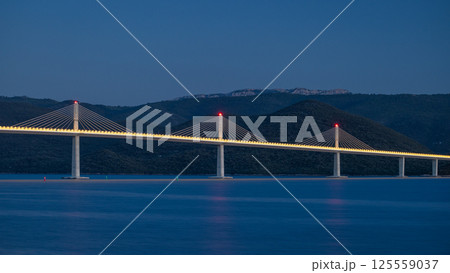 The Peljesac Bridge to Peljesac peninsula in Croatia, Europe. A modern cable-stayed bridge illuminated against a blue evening sky, spanning over calm waters with mountains in the background. 125559037