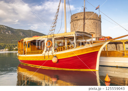 Mali Ston, village in Peljesac peninsula in Croatia, Europe. Colorful wooden boat reflecting in calm water near a stone tower under a partly cloudy sky in a coastal scene. 125559043