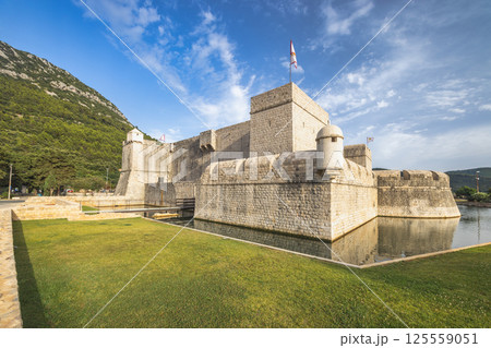 Ston, historic town in Peljesac peninsula in Croatia, Europe. Stone fortress with a moat and flag under a blue sky, against a green mountain backdrop. Tranquil waters reflect the fort. 125559051