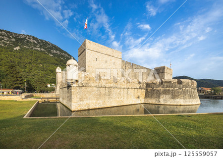 Ston, historic town in Peljesac peninsula in Croatia, Europe. Fortified walls and moat of an ancient European castle, with lush green backdrop and brilliant blue sky. A stately fortress. Ston, historic town in Peljesac peninsula in Croatia, Europe. Fortified walls and moat of an ancient European castle, with lush green backdrop and brilliant blue sky. A stately fortress. 125559057