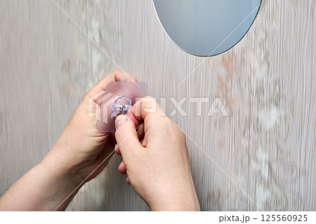 Hands are removing protective film from nano adhesive hook before mounting it onto smooth plastic wall panel near round mirror. 125560925