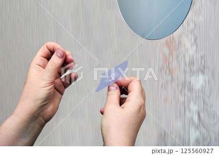 Hands are peeling off protective film from peel-and-stick hook before attaching it to smooth plastic wall panel near circular mirror. Hands are peeling off protective film from peel-and-stick hook before attaching it to smooth plastic wall panel near circular mirror. 125560927