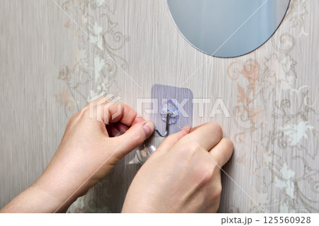 Person is pressing transparent adhesive patch with plastic hook onto vertical surface near round mirror, using both hands for secure attachment. Person is pressing transparent adhesive patch with plastic hook onto vertical surface near round mirror, using both hands for secure attachment. 125560928
