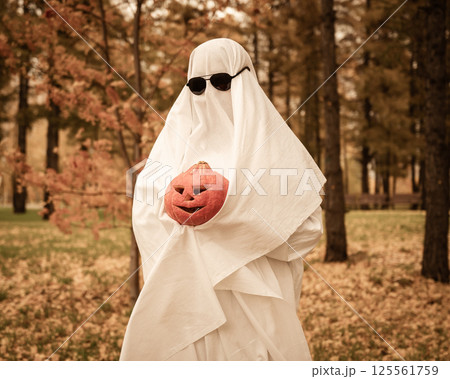 Woman in white sheet holding jack o'lantern against autumn forest background. Halloween costume. Woman in white sheet holding jack o'lantern against autumn forest background. Halloween costume. 125561759