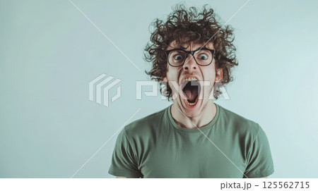 Portrait of a furious young man with curly hair and eyeglasses shouting and showing negative emotions against a light blue background, expressing anger and frustration 125562715
