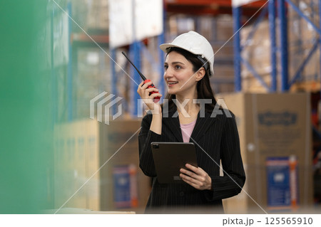 Female worker uses walkie talkie digital tablet checking stock goods working in logistics. Women professional warehouse worker wearing safety uniform and white hard hat inspect product at storage. 125565910