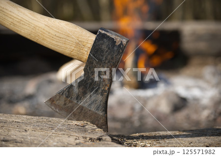 An axe is deeply embedded in a wood chopping block with a warm campfire blazing behind it. 125571982