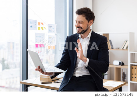 Businessman with beard in suit sitting in modern office having online conference waving, engaging in communication through laptop. Positive and professional workplace environment showcasing technology 125572149