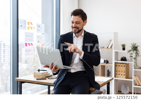 Professional adult Caucasian man wearing suit seated at desk holding laptop explaining concept 125572150