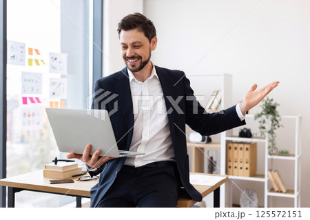 Young adult male business professional wearing suit sitting at desk conducting video meeting. Workplace setting includes books and plants. Confidence and enthusiasm displayed through gestures 125572151