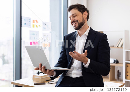 Professional adult Caucasian male in formal suit on video call speaking and gesturing at laptop, in bright modern office setting. Captures positive communication, digital technology, business concept. Professional adult Caucasian male in formal suit on video call speaking and gesturing at laptop, in bright modern office setting. Captures positive communication, digital technology, business concept. 125572159