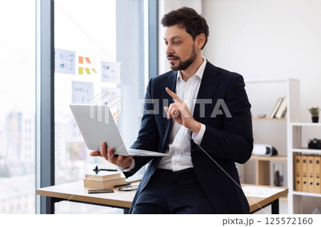 Professional adult Caucasian man wearing suit seated at desk holding laptop explaining concept Professional adult Caucasian man wearing suit seated at desk holding laptop explaining concept 125572160