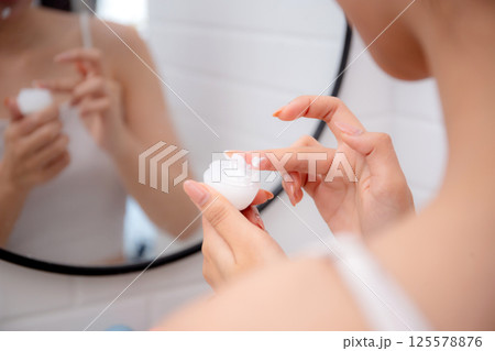 Closeup hand of woman applying cream or lotion for rejuvenation skin on face while looking mirror. 125578876