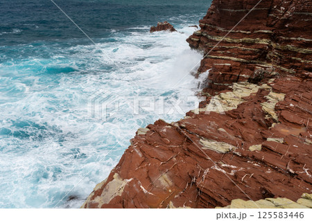 Red rock cliff in Cala Rossa, Terrasini, coastal waves crashing against rugged cliffs during daylight Red rock cliff in Cala Rossa, Terrasini, coastal waves crashing against rugged cliffs during daylight 125583446