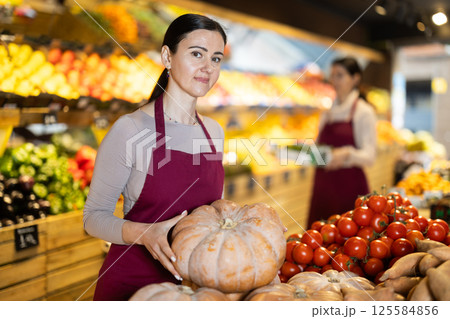 Adult woman seller puts pumpkin in vegetable shop Adult woman seller puts pumpkin in vegetable shop 125584856