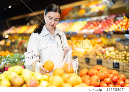 Focused positive middle aged woman shopping in organic food store, choosing oranges 125585074