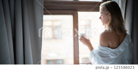 Woman in elegant robe drinking coffee in hotel room and standing near window 125586023