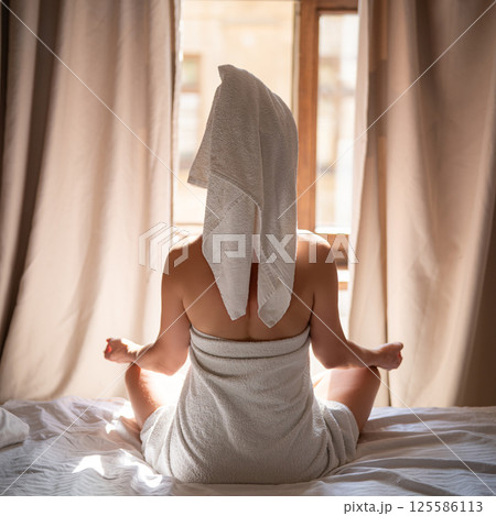 Back view of young woman in towel sitting on bed and meditating 125586113