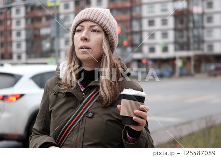 Portrait of mature woman in wool hat standing on the urban street, drinking takeaway coffee from paper cup. Middle-aged 40s female walking around the city at cold autumn day. Living in modern district Portrait of mature woman in wool hat standing on the urban street, drinking takeaway coffee from paper cup. Middle-aged 40s female walking around the city at cold autumn day. Living in modern district 125587089