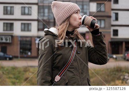 Portrait of mature woman in wool hat standing on the urban street, drinking takeaway coffee from paper cup. Middle-aged 40s female walking around the city at cold autumn day. Living in modern district Portrait of mature woman in wool hat standing on the urban street, drinking takeaway coffee from paper cup. Middle-aged 40s female walking around the city at cold autumn day. Living in modern district 125587126