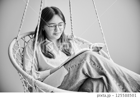 Beautiful girl relaxing and reading book on indoor swing. Pretty female in glasses excited about story she reading. Cozy afternoon with book in hanging chair in home. Black and white image. Beautiful girl relaxing and reading book on indoor swing. Pretty female in glasses excited about story she reading. Cozy afternoon with book in hanging chair in home. Black and white image. 125588501