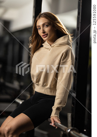 fit woman wearing beige hoodie and black shorts poses playfully on gym equipment. bright environment highlights her focused demeanor and dedication fitness. fit woman wearing beige hoodie and black shorts poses playfully on gym equipment. bright environment highlights her focused demeanor and dedication fitness. 125591080