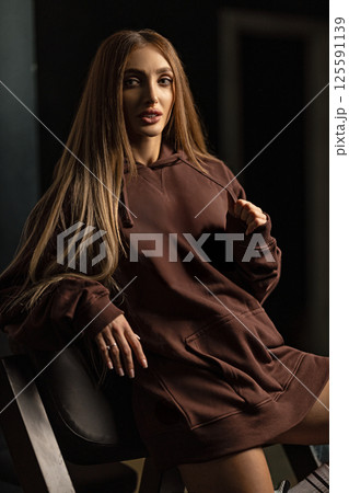 woman long hair poses confidently while seated on chair dressed in brown hoodie. studio's soft lighting highlights her features and creates intriguing atmosphere. woman long hair poses confidently while seated on chair dressed in brown hoodie. studio's soft lighting highlights her features and creates intriguing atmosphere. 125591139