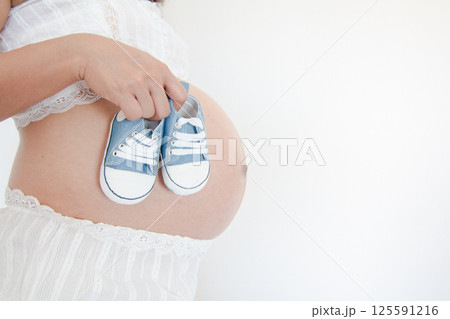 Mother-to-Be Holding Baby Shoes with Anticipation Against Bare Belly 125591216