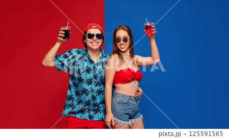 Couple in beachwear holding drinks, smiling to camera with confident summer vibe against split red blue background. Couple in beachwear holding drinks, smiling to camera with confident summer vibe against split red blue background. 125591565