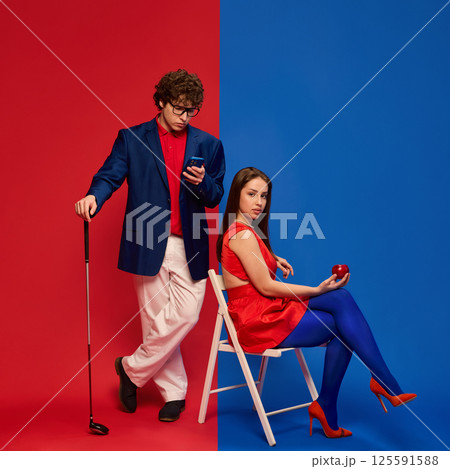Couple, young man and woman in matching color outfits, with golf club and apple, showing quirky elegance against split blue and red background. 125591588