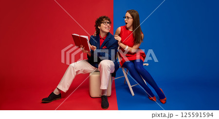 Man and woman with excited and shocked faces reading book, sharing joyful moment against split red and blue background. 125591594