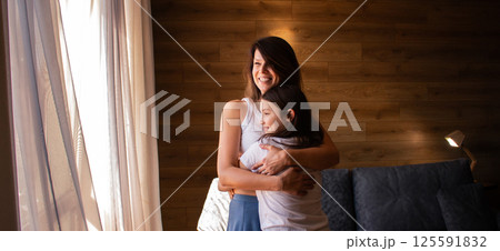Portrait of Caucasian mom and daughter hug and cuddle in living room standing by window. Understanding relationships, love and family bonding concept. 125591832