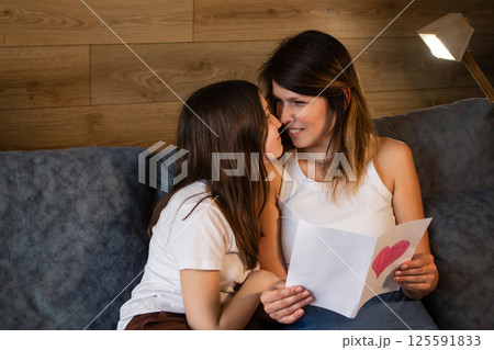 Mom and her preteen daughter reading together greeting card with drawn heart sitting on sofa at home. Spending quality time with children. 125591833