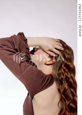 young woman with long wavy red hair touching her head with hand in stylish brown dress with open back against light background, beauty salon, haircare, fashion industry 125591962