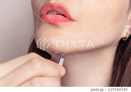 A woman's beard grows. A woman pulls dark hair from her chin with tweezers. Close up cropped photo A woman's beard grows. A woman pulls dark hair from her chin with tweezers. Close up cropped photo 125592585