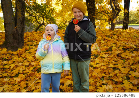 A cheerful boy and girl enjoy colorful lollipops in a scenic autumn park, surrounded by golden leaves. Dressed in cozy fall clothing, they smile playfully, capturing the warmth and joy of the season. A cheerful boy and girl enjoy colorful lollipops in a scenic autumn park, surrounded by golden leaves. Dressed in cozy fall clothing, they smile playfully, capturing the warmth and joy of the season. 125596499