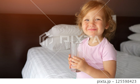 Little girl drinks water from clear glass perched on bed edge 125598423