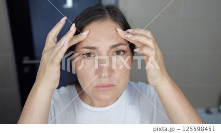 Woman stands at bathroom mirror carefully examining face 125598451
