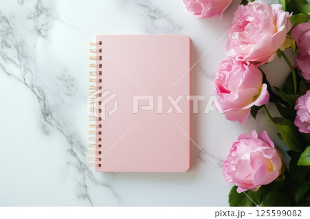 Soft pink notebook with spiral binding placed on elegant marble surface. Beautiful blooming peonies on the side create romantic and gentle flat lay. Ideal for feminine design or planning themes. 125599082