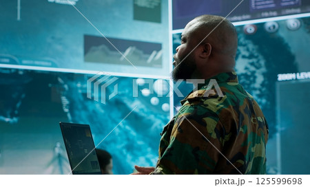 Military officer oversees surveillance and cybersecurity operations in a high tech army command post, ensuring the protection of classified information and data integrity. Big screen gear. Camera A. Military officer oversees surveillance and cybersecurity operations in a high tech army command post, ensuring the protection of classified information and data integrity. Big screen gear. Camera A. 125599698