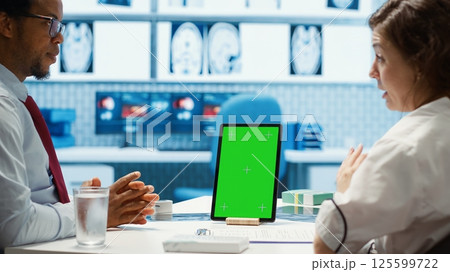 General practitioner pointing at green screen mockup on device, discussing with her patient about recovery treatment. Doctor doing checkup visit on insurance with a man. Camera A 125599722