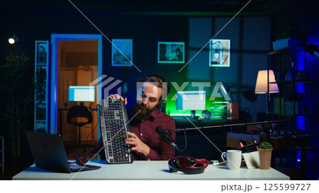 Man in studio reviewing gaming keyboard received from sponsoring brand. Online show host paid to promote bluetooth wireless computer peripheral to viewers during live broadcast, camera B Man in studio reviewing gaming keyboard received from sponsoring brand. Online show host paid to promote bluetooth wireless computer peripheral to viewers during live broadcast, camera B 125599727