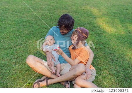 Father with his two sons in the park, the older boy wearing a pilot hat and holding a toy airplane while the baby watches. Childhood, bonding, and family adventure concept Father with his two sons in the park, the older boy wearing a pilot hat and holding a toy airplane while the baby watches. Childhood, bonding, and family adventure concept 125600371