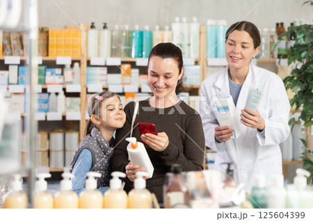 Woman with daughter in pharmacy scans QR code on cream, pharmacist in background 125604399