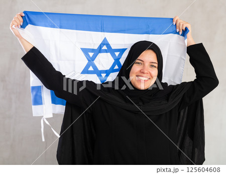 Balanced smiling Muslim woman in traditional black hijab holds flag of Israel Balanced smiling Muslim woman in traditional black hijab holds flag of Israel 125604608